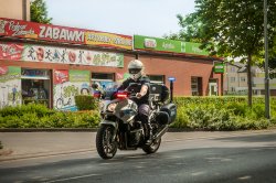 Policjant Ruchu Drogowego w umundurowaniu - kombinezonie motocyklowym jedzie ulicą motorem służbowym z włączonymi sygnałami uprzywilejowania.