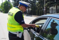 Policjant służby ruchu drogowego ubrany w mundur służbowy i kamizelkę odblaskową prowadzi badanie stanu trzeźwości kierowcy samochodu osobowego urządzeniem alcobloe do wstępnego badania.