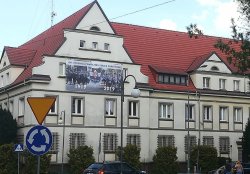 Baner na budynku Komendy Powiatowej Policji w Złotowie z okazji 100. rocznicy powołania Policji Państwowej. Baner Przedstawia po lewej policjantów pełniących służbę w 1919 roku i teraźniejszych funkcjonariuszy po prawej stronie baneru