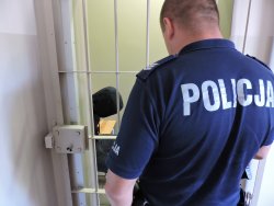 policjant w umundurowaniu stoi przed pomieszczeniem dla osób zatrzymanych. Pilnując zatrzymanego mężczyznę który siedzi na ławce za kratą. Na głowie ma założony kaptur