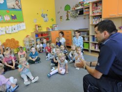 Dzielnicowy aspirant Jacek Kozdra rozmawia z przedszkolakami na temat bezpieczeństwa w ruchu drogowym. Policjant umundurowany siedzi na krześle dzieci zaś siedzą na dywanie w półkolu wsłuchując się w słowa policjanta.