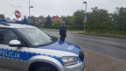 Policjantka ruchu drogowego sprawuje nadzór nad przejściem dla pieszych. Policjantka w umundurowaniu służbowym stoi obok oznakowanego radiowozu