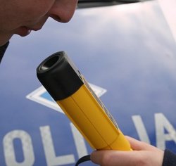 Policjant prowadzi badanie stanu trzeźwości kierującego urządzeniem alcooblowe. W tle policyjny radiowóz.