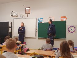 Dzielnicowa sierżant sztabowy Monika Drabowicz i starszy dzielnicowy aspirant Jacek Kozdra prowadzą pogadankę dla uczniów klasy trzeciej. Policjanci stoją przy tablicy w klasie lekcyjnej , dzieci zaś przysłuchują się poruszanym tematom siedząc w ławkach.