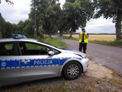 Policjant ruchu drogowego stoi na poboczu drogi i dokonuje pomiaru prędkości przy użyciu ręcznego miernika prędkości. Policjant stoi obok ustawionego prostopadle do jezdni radiowozu oznakowanego.