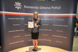 Sierżant Monika Kapera stoi na tle logo Komendy Głównej Policji w umundurowaniu galowym trzymając w ręce wyróżnienie