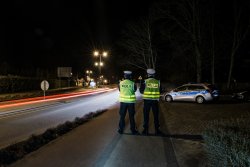 Policjanci ruchu drogowego stoją odwróceni tyłem do jezdni obserwując ruch odbywający się na drodze. Po prawej stronie widoczny policyjny radiowóz oznakowany.