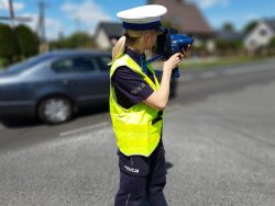 Policjantka prowadzi kontrolę ruchu drogowego. Dokonuje pomiaru prędkości laserowym miernikiem stojąc przy drodze w tle widać mijający policjantkę samochód