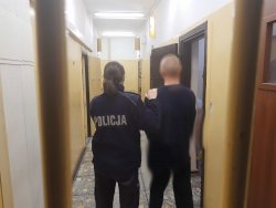 Zatrzymany mężczyzna stoi tyłem po lewej stronie stoi umundurowana policjantka trzymająca zatrzymanego mężczyznę. W  tle widać korytarz pomieszczenia dla osób zatrzymanych