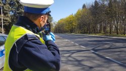 Policjant prowadzi pomiar prędkości pojazdów przy użyciu ręcznego miernika prędkości.Widać drogę i stojącego po lewej stronie policjanta służby ruchu drogowego i w tle policyjny radiowóz oznakowany