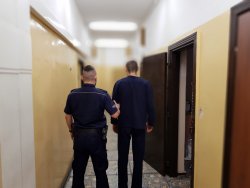 Zatrzymany mężczyzna stoi ubrany w niebieski dres po lewej stronie stoi policjant w umundurowaniu służbowym i trzyma zatrzymanego 30-latka