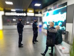 Policjanci kontrolują galerię handlową rozmawiają z osobami przebywającymi w jej wnętrzu. Policjanci stoją a jedna z osób siedzi na ławce druga zaś stoi