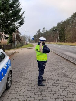 Policjantka Wydziału Ruchu Drogowego prowadzi pomiar prędkości przy użyciu ręcznego miernika prędkości stoi przy drodze po lewej stronie obok ustawiony jest policyjny radiowóz oznakowany. W tle widać nadjeżdżający samochód