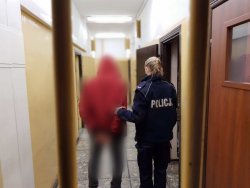 Policjantka trzyma zatrzymanego mężczyznę który ma założone kajdanki na ręce trzymane z tyłu. Fotografia wykonana w pomieszczeniu dla osób zatrzymanych tło stanowią kraty pomieszczenia