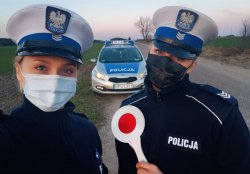 Policjantki ruchu drogowego wykonały sobie zdjęcie tzw. selfie stoją w umundurowaniu z maseczkami policjantka po prawej stronie trzyma tarczę do zatrzymywania pojazdów. W tle widoczny radiowóz oznakowany