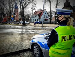 Policjantka ruchu drogowego obserwuje przejście dla pieszych stojąc przy policyjnym radiowozie oznakowanym