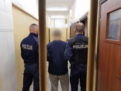 Policjanci w umundurowaniu służbowym trzymają zatrzymanego mężczyznę, który stoi w pomieszczeniu dla osób zatrzymanych. Jeden policjant stoi po lewej stronie zatrzymanego drugi zaś po prawej stronie. W tle widać karaty pomieszczenia.