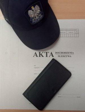 Na zdjęciu widać telefon komórkowy w etui oraz czapkę policyjną leżącą na aktach