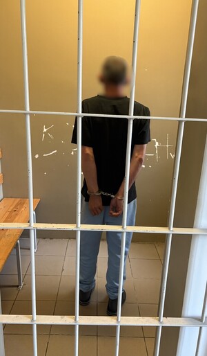 Zatrzymany mężczyzna, z kajdankami na rękach trzymanych z tyłu, stojący w pomieszczeniu za kratą